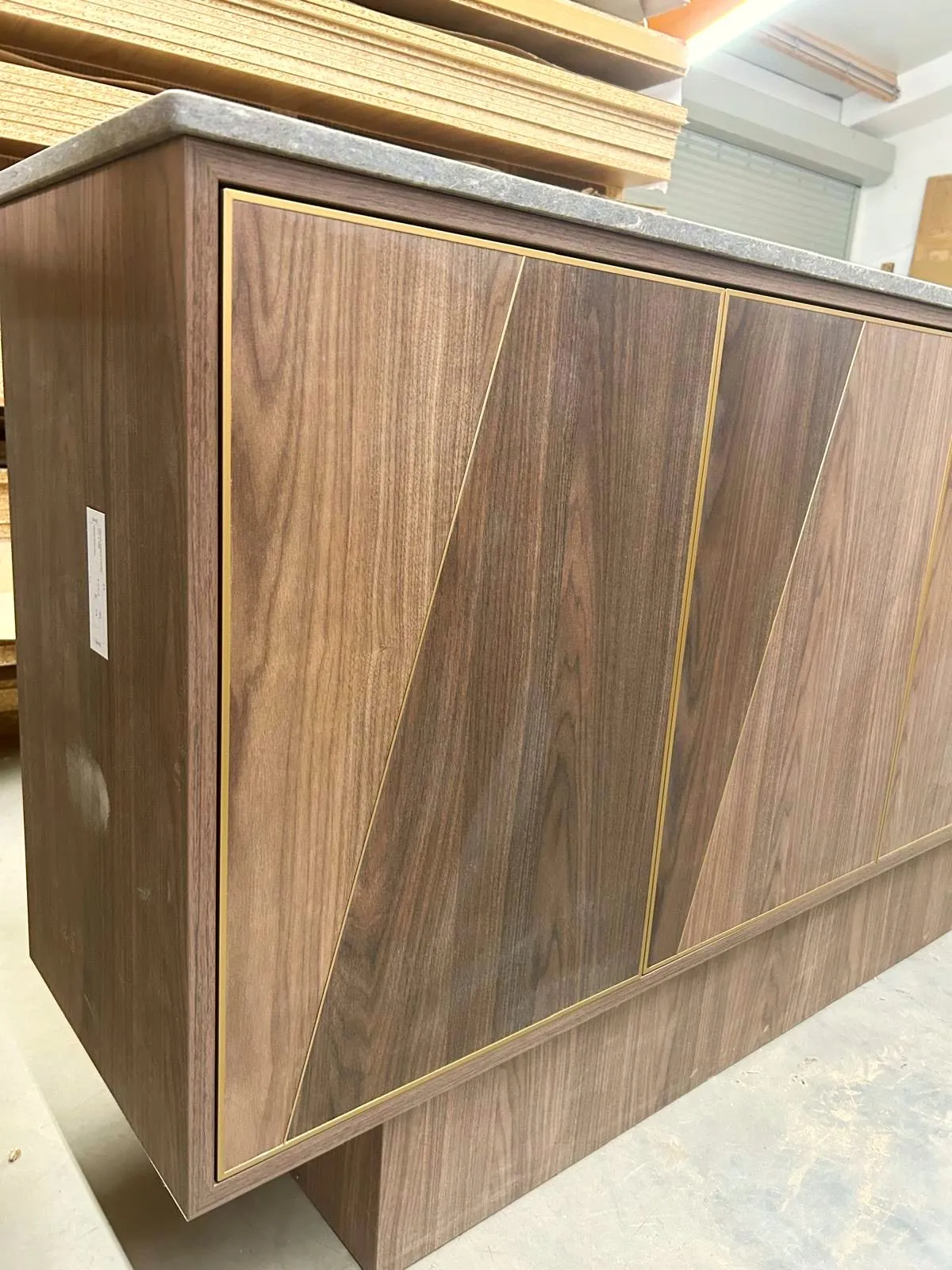 Nahaufnahme einer Schrankecke mit diagonaler Holzmaserung, Messingintarsien und Natursteinplatte in der Werkstatthalle